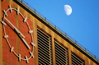 Blick auf die Uhr am Kirchturm, rechts seitlich ist der Halbmond vor blauem Himmel zu sehen