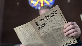 Ein Mann liest in einer alten Gemeindezeitung. Im Hintergrund ist das große Rundfenster verschwommen zu sehen.