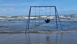 Eine Schaukel am Strand, die vom Meer umspült wird. Ein Mann schaukelt.