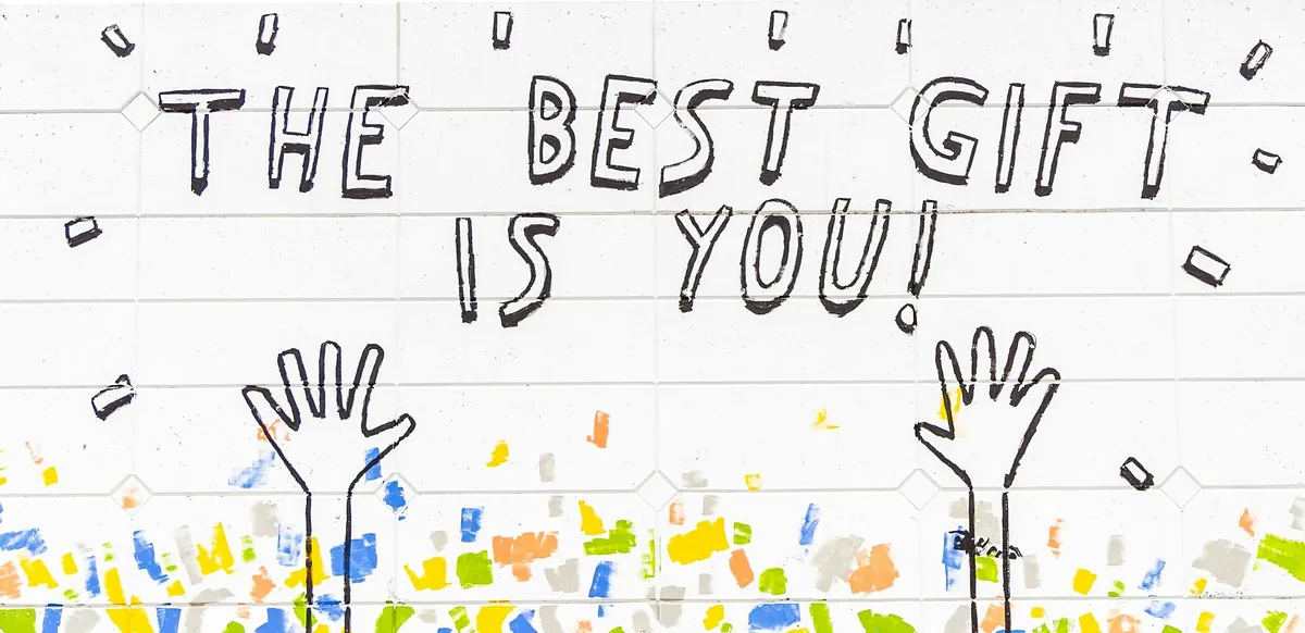 Eine weiße Wand mit bunten Flecken, zwei gezeichneten Händen und dem Satz "The best gift ist you!"